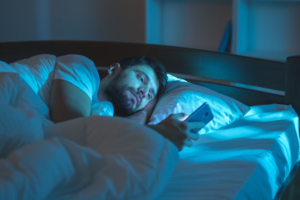 Man is alone in bed reading his phone things you're doing that would horrify sleep doctors