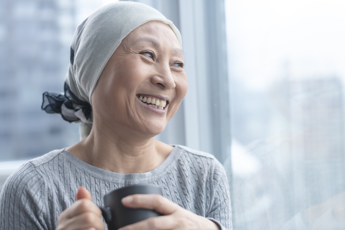 A Korean senior woman with cancer is wearing a scarf on her head. She is standing and holding a cup of tea. The woman leans against a window and smiles with gratitude and hope.