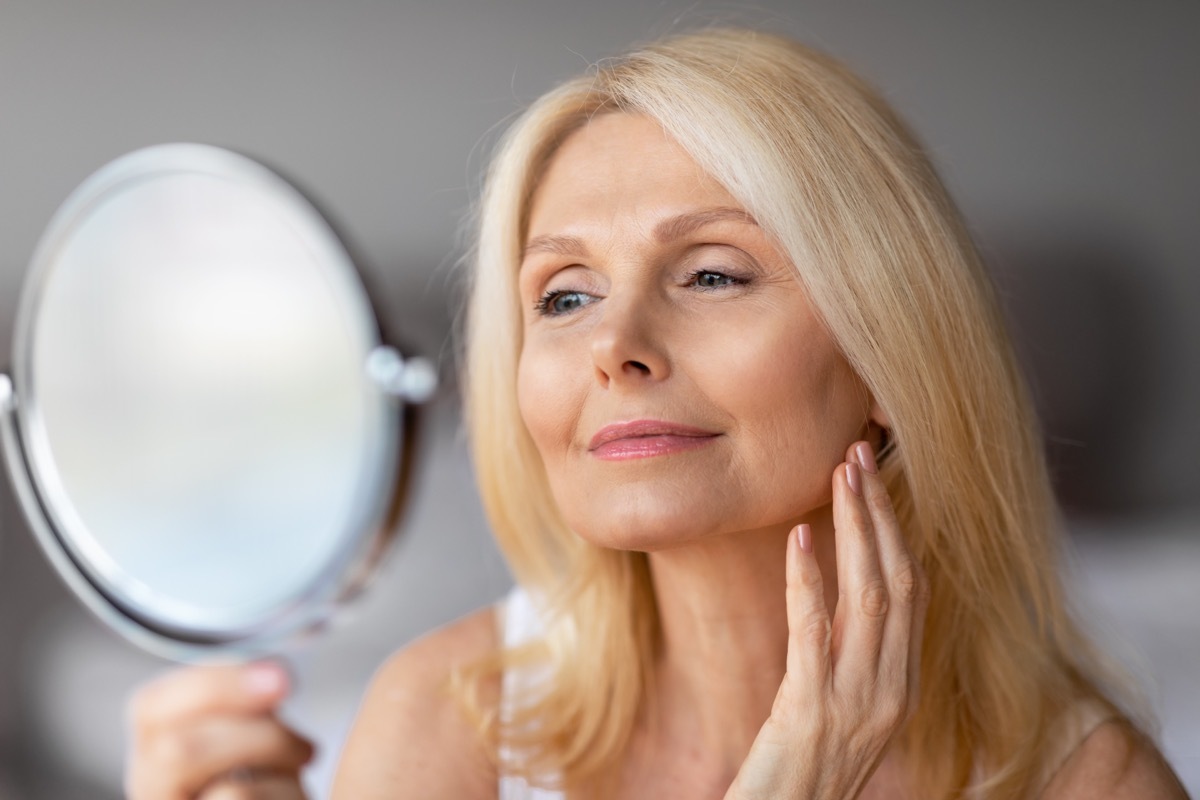 Attractive aged european woman looking in magnifying mirror and touching her face smooth skin, beautiful senior lady enjoying her appearance, closeup