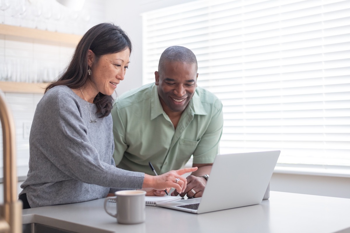 couple use a laptop computer to pay bills online, manage budget and prepare tax documents. They are in the kitchen of their home.