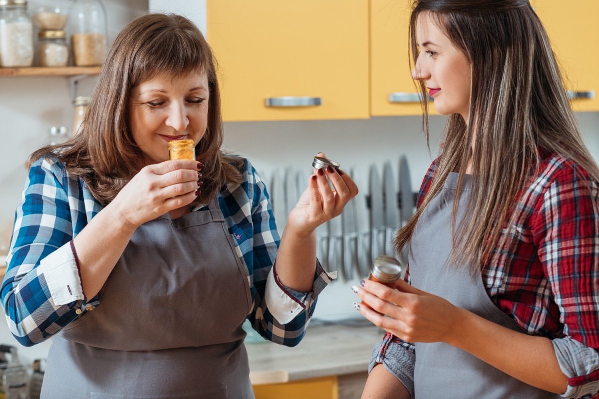 Two women in kitchen cooking. Mother smelling spices with smile. Selecting choosing ingredients. Daughter look waiting.