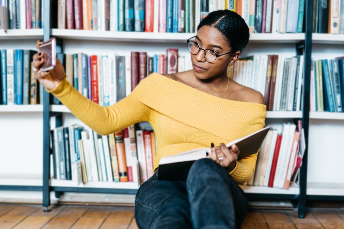 african american young woman student in eyewear making selfie photo on front camera of smartphone for sharing in networks sitting with literature book on floor and preparing for exam in library