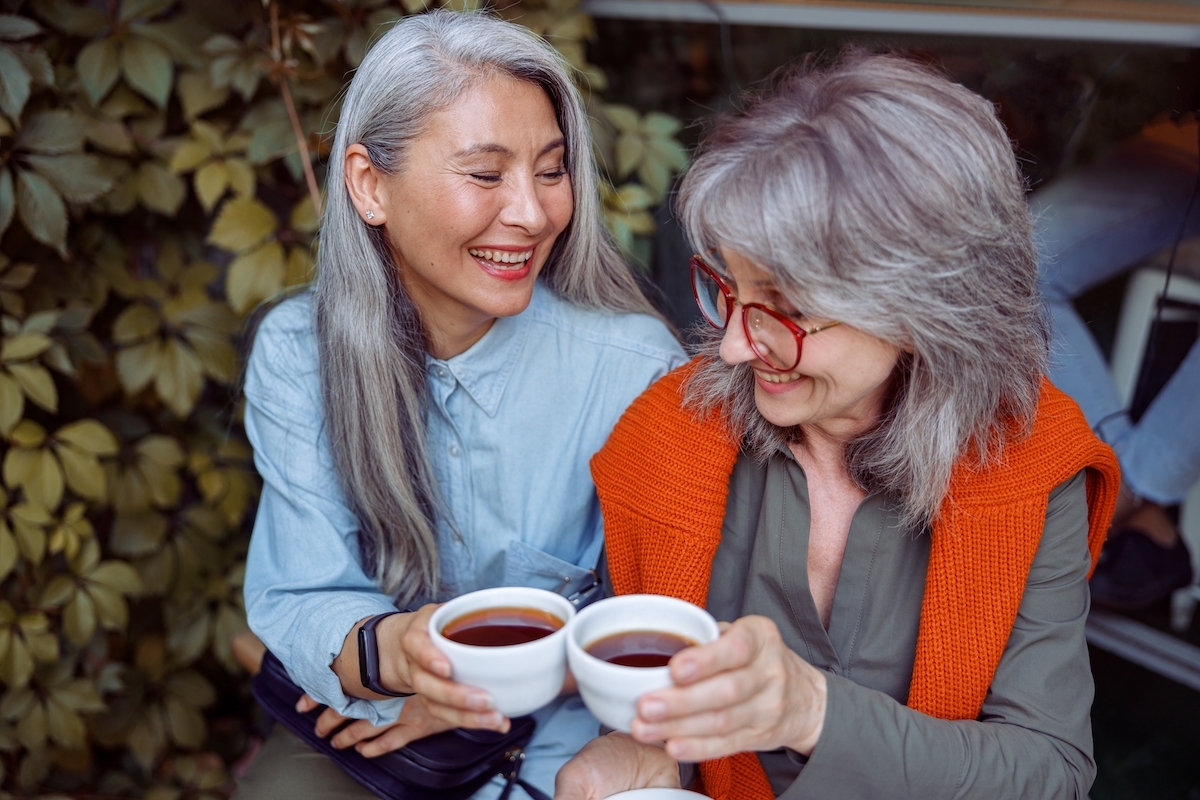 Two women friends with long gray hair having coffee and smiling.