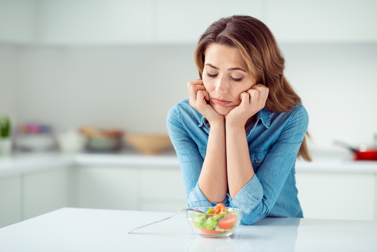 middle aged woman looks disappointed at small salad while standing in kitchen