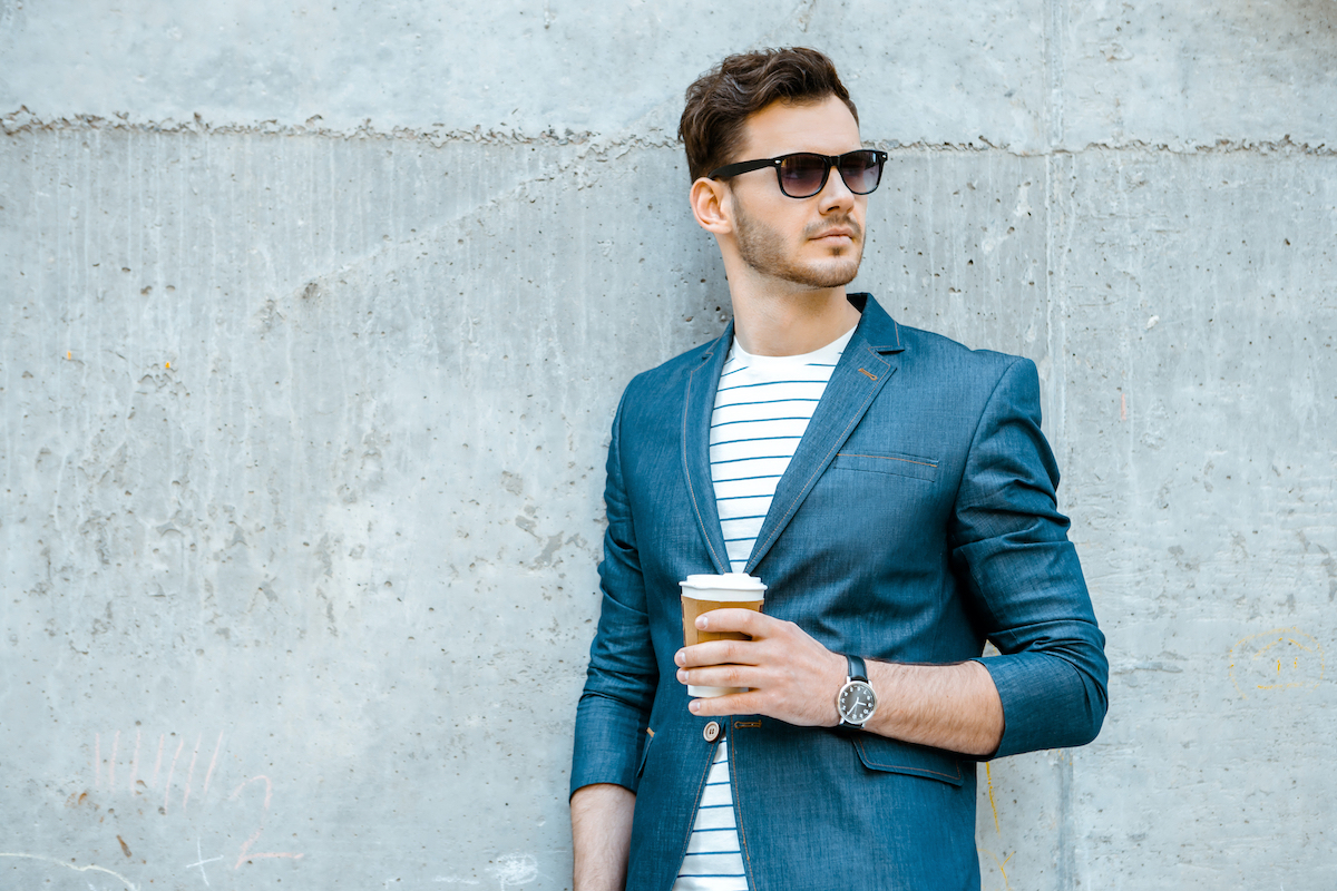 Portrait of stylish handsome young man with bristle standing outdoors and leaning on wall. Man wearing jacket, sunglasses, shirt and holding cup of coffee