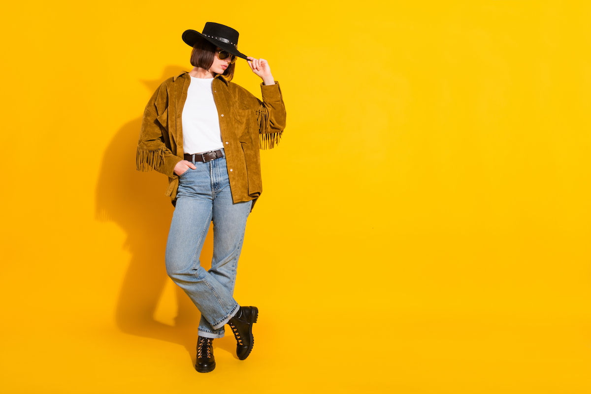 Young woman wearing a black cowboy hat, a brown suede fringe jacket, jeans, and black boots—against a yellow background.