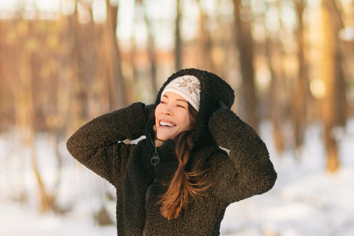 A young, smiling woman wearing a black sherpa jacket stands outside on a snowy day.