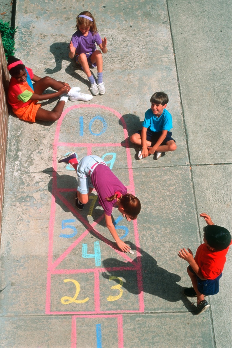 1980s GROUP OF FIVE CHILDREN BOYS AND GIRLS PLAYING HOPSCOTCH ON CONCRETE SIDEWALK