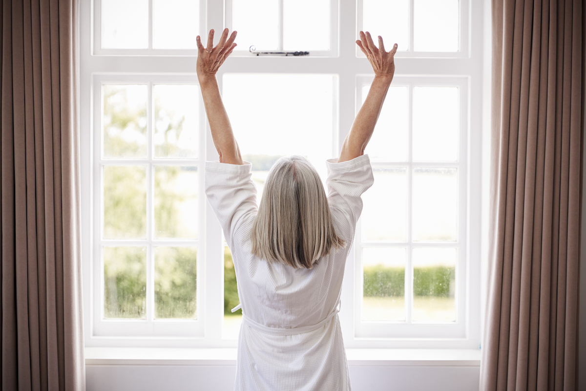 Rear view of a woman with gray hair wearing a white bathrobe stretching in the morning in front of a window