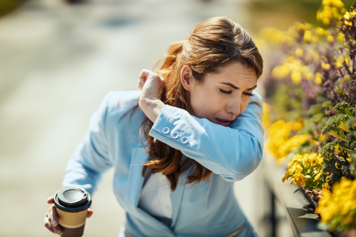 woman coughing into her elbow outside while holding coffee