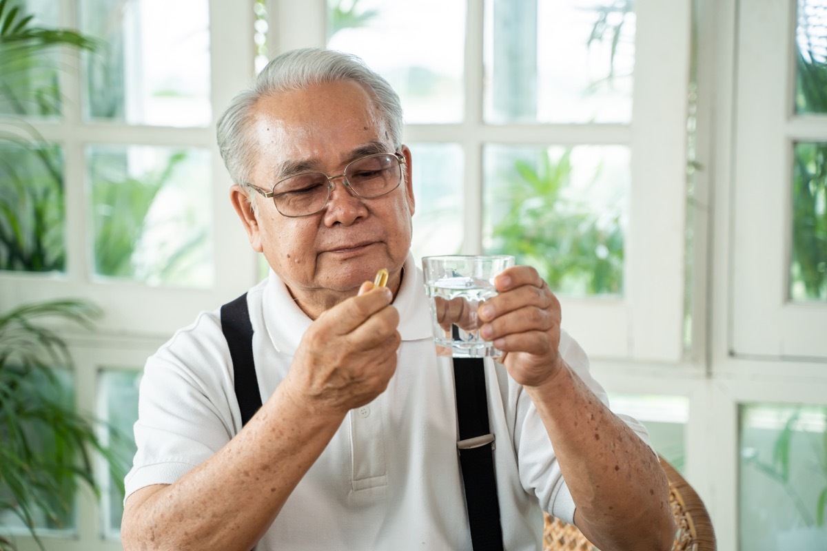  man with cup of water and pill looking at camera and taking remedy in cozy room at home. Old male taking medicine.