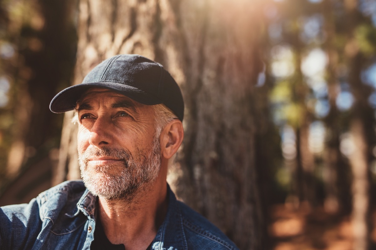 older white man in a black hat sitting in front of a giant tree and looking peaceful