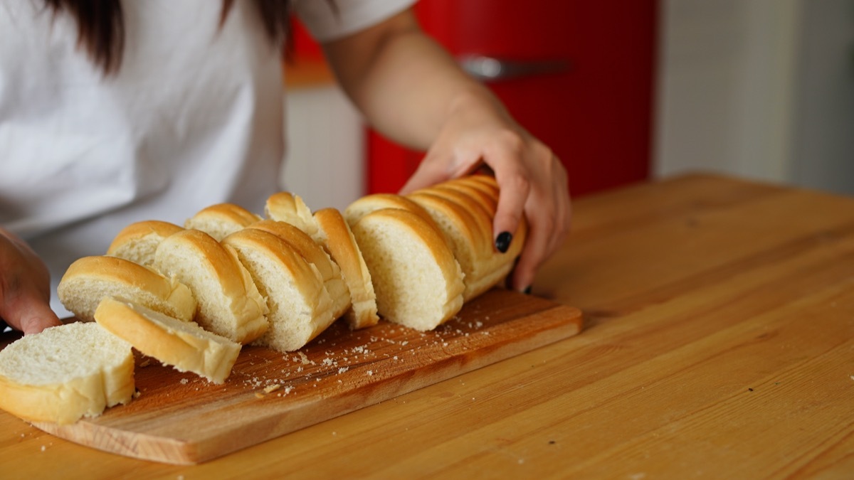 Women's hands cutting baguette with knife on chopping board on kitchen table.