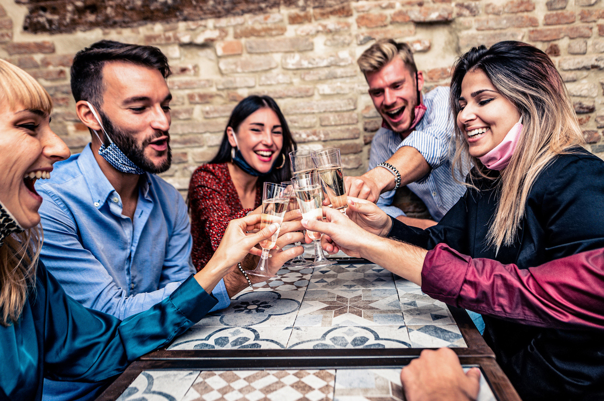 Young people with face masks on chins toasting with champagne flutes amid coronavirus