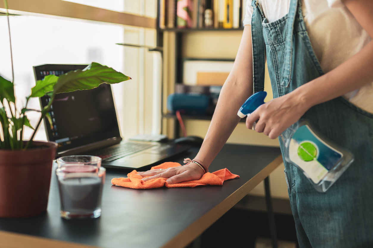 A woman cleans a desk with a laptop on it using a spray bottle with disinfectant and an orange rag.