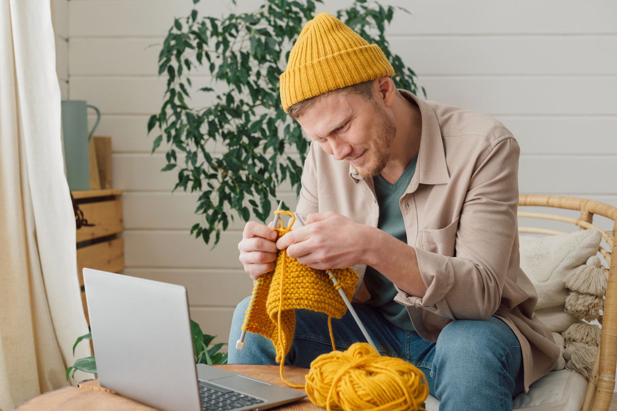 A man watching a video to learn how to knit a yellow scarf to match his yellow hat.