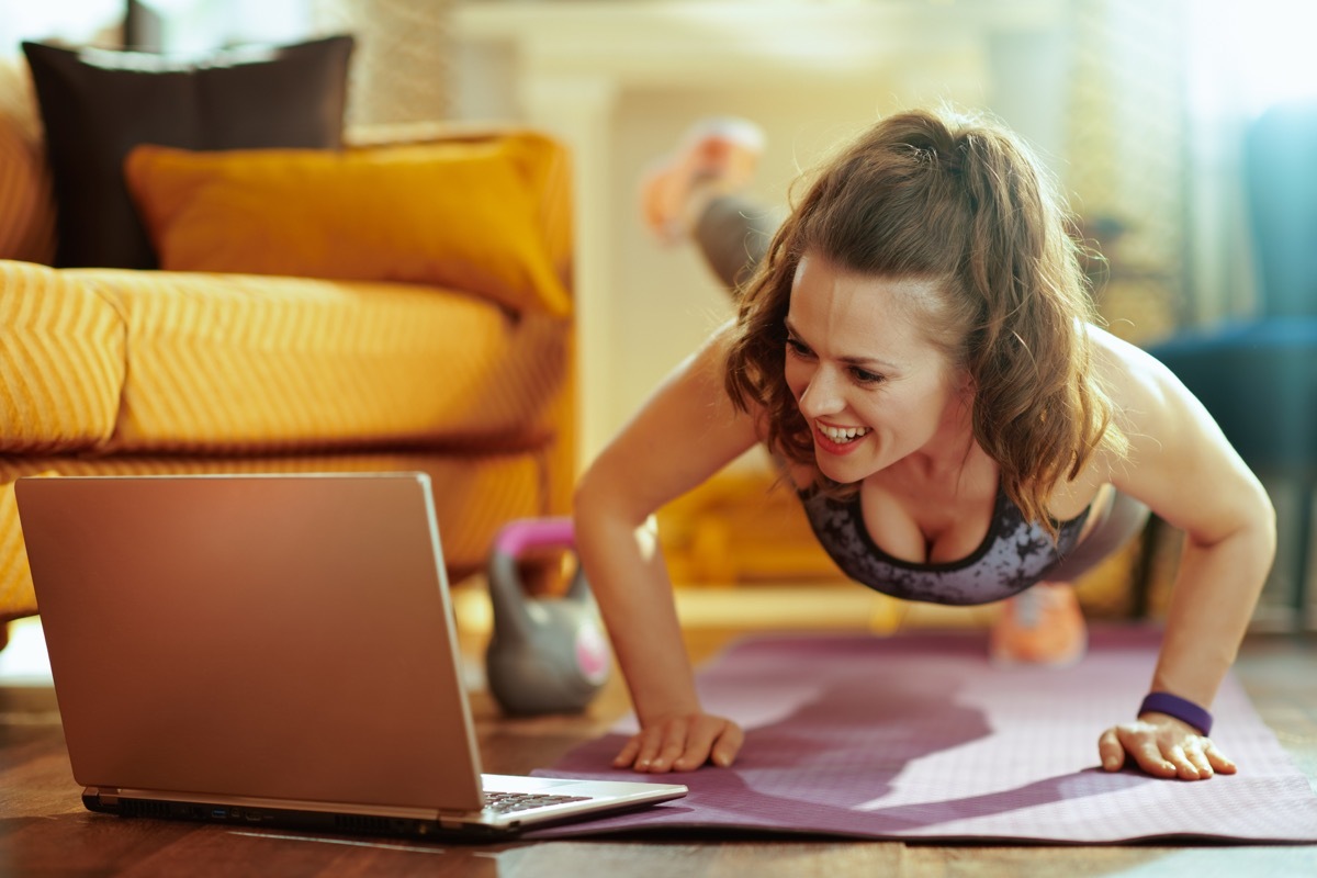 smiling healthy woman in fitness clothes in the modern living room watching fitness tutorial on internet via laptop and doing pushups on fitness mat. (smiling healthy woman in fitness clothes in the modern living room watching fitness tutorial on inte