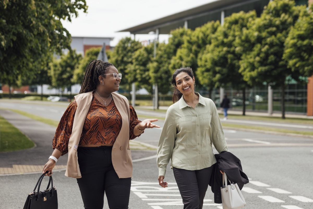 A three quarter length shot of two businesswomen walking and talking to each other after work. They are both dressed smartly and casually and are walking over a road. They are based in the North East of England.