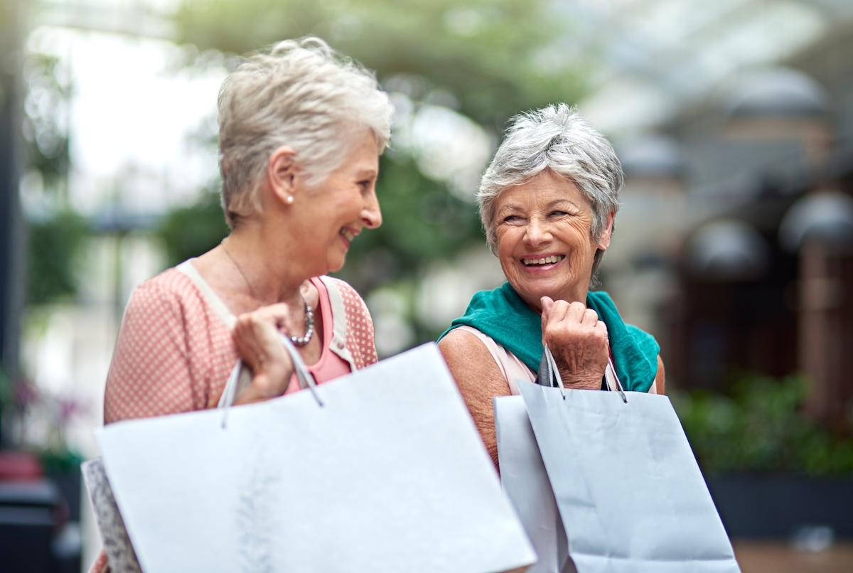 Two senior female friends holding shopping bags and smiling at each other.