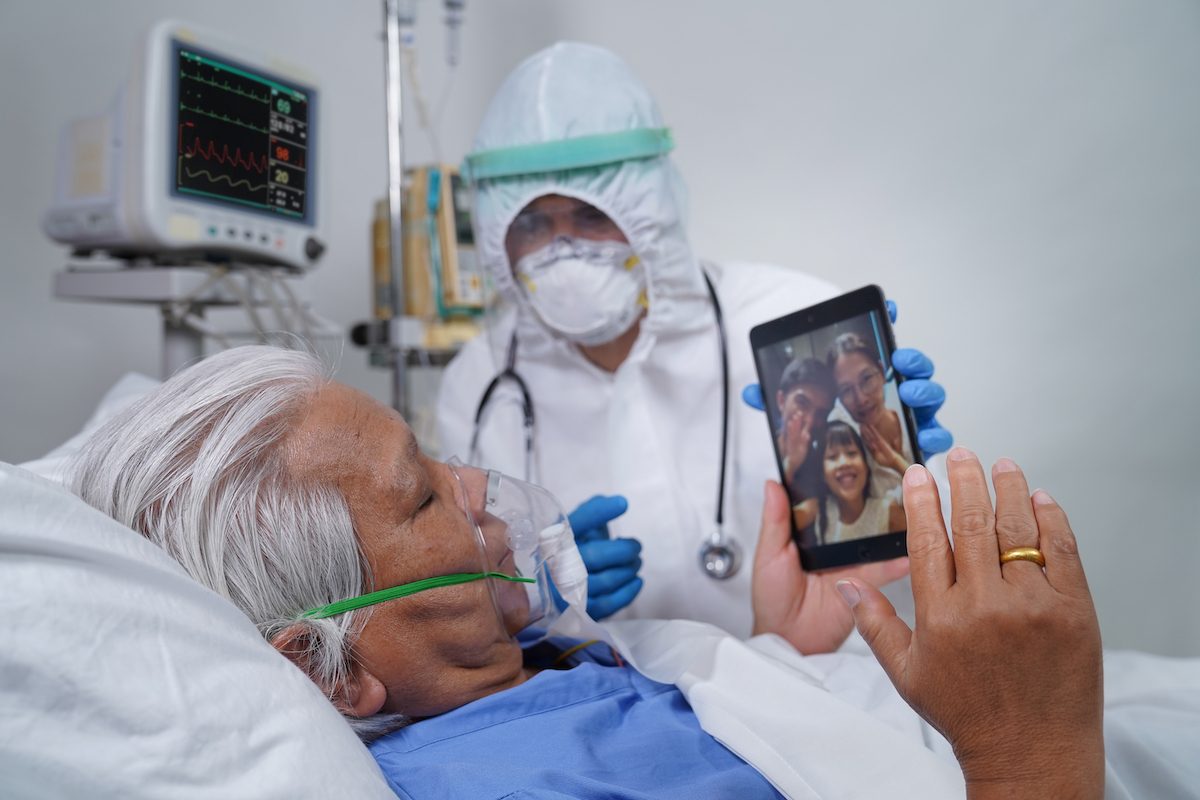 Doctor helps elderly coronavirus patient in the hospital, communicating with his family using tablet video call.