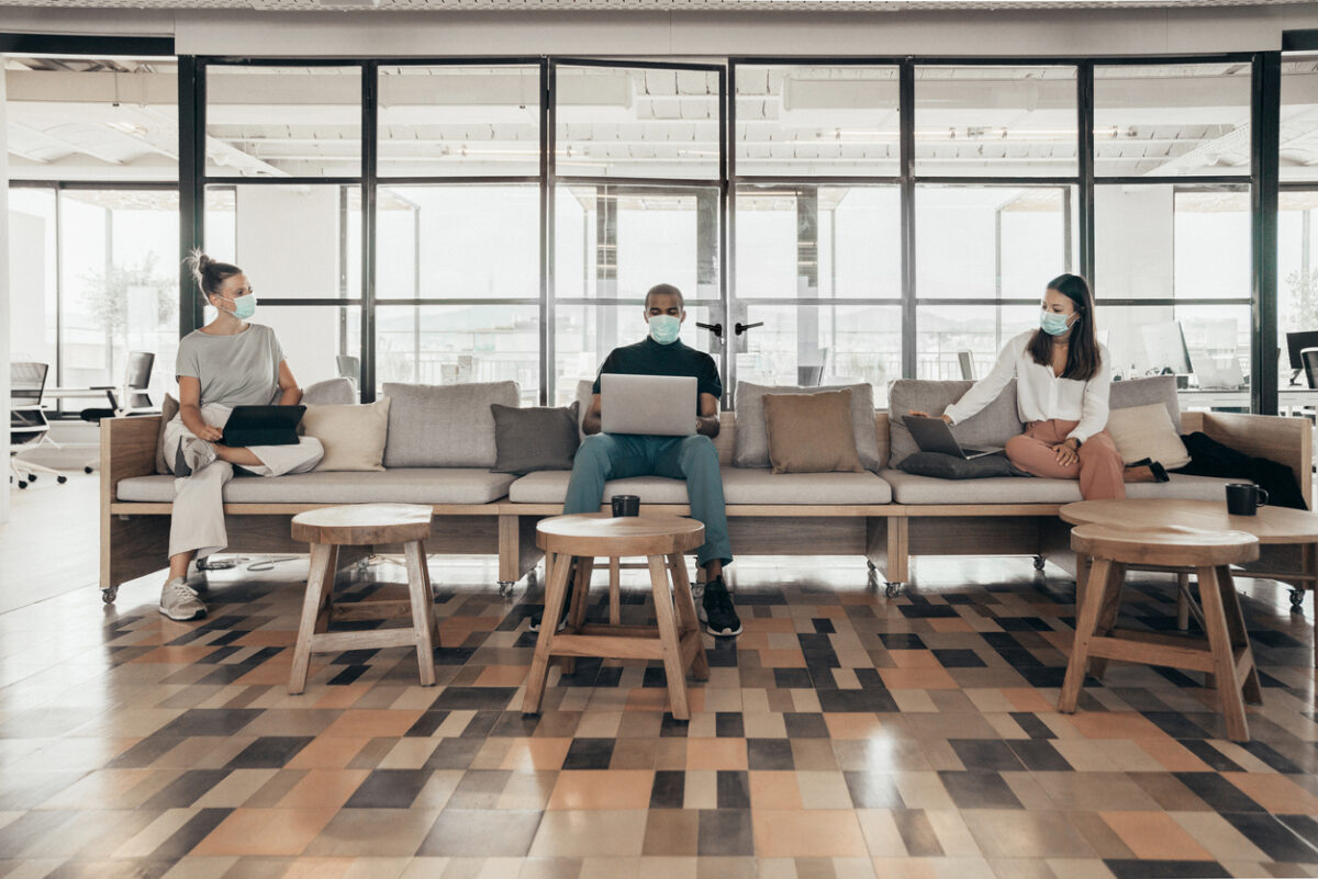 Three coworkers sit about six feet apart from each other on a couch in their office.