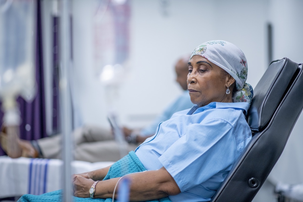 A woman wearing a head scarf lies on a hospital bed and looks to the side in contemplation. She is wearing a head scarf and a hospital gown and there is a IV drip next to her.