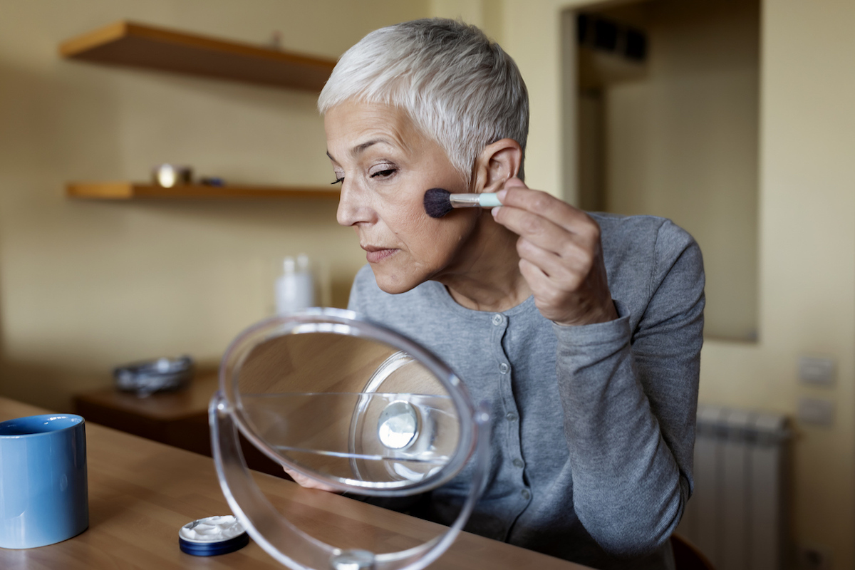 A woman with short gray hair wearing a gray cardigan puts makeup on in a small mirror.