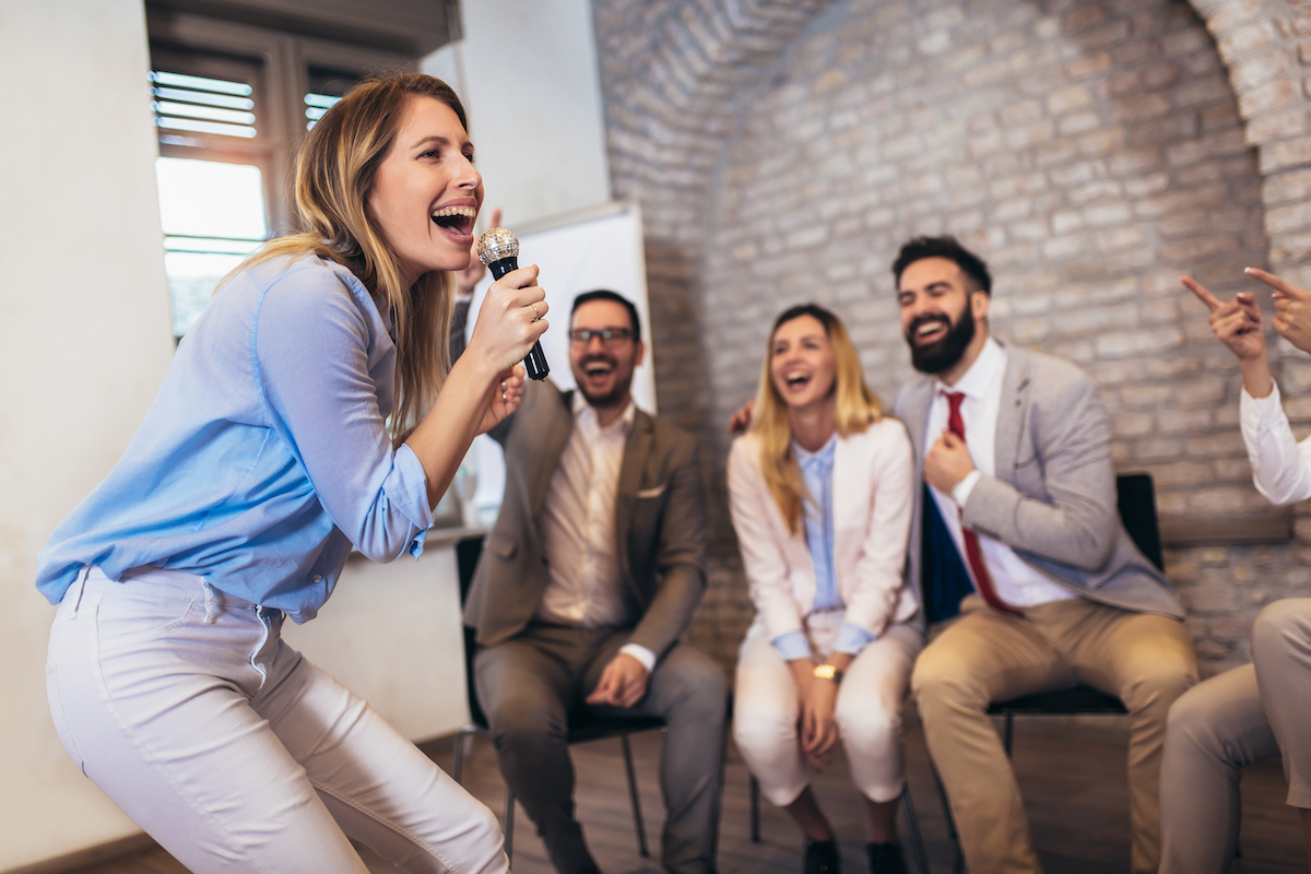 Close up of a young blonde woman singing karaoke with her coworkers sitting behind her.