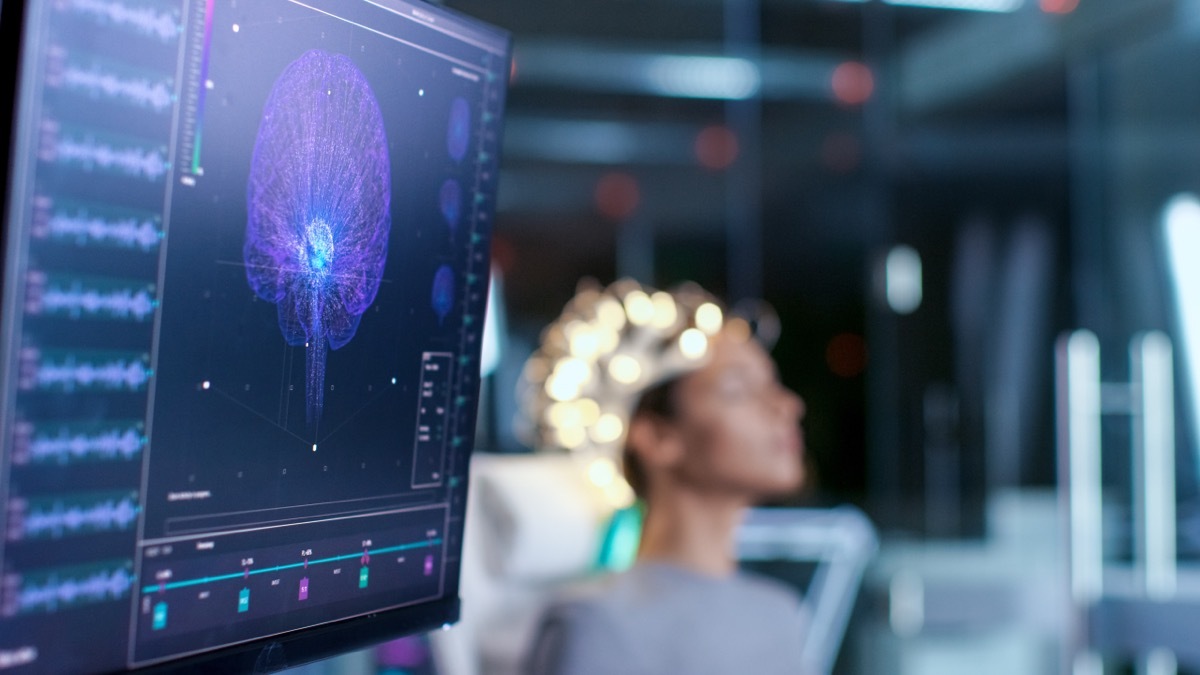 Woman Wearing Brainwave Scanning Headset Sits in a Chair In the Modern Brain Study Laboratory/ Neurological Research Center. Monitors Show EEG Reading and Brain Model.