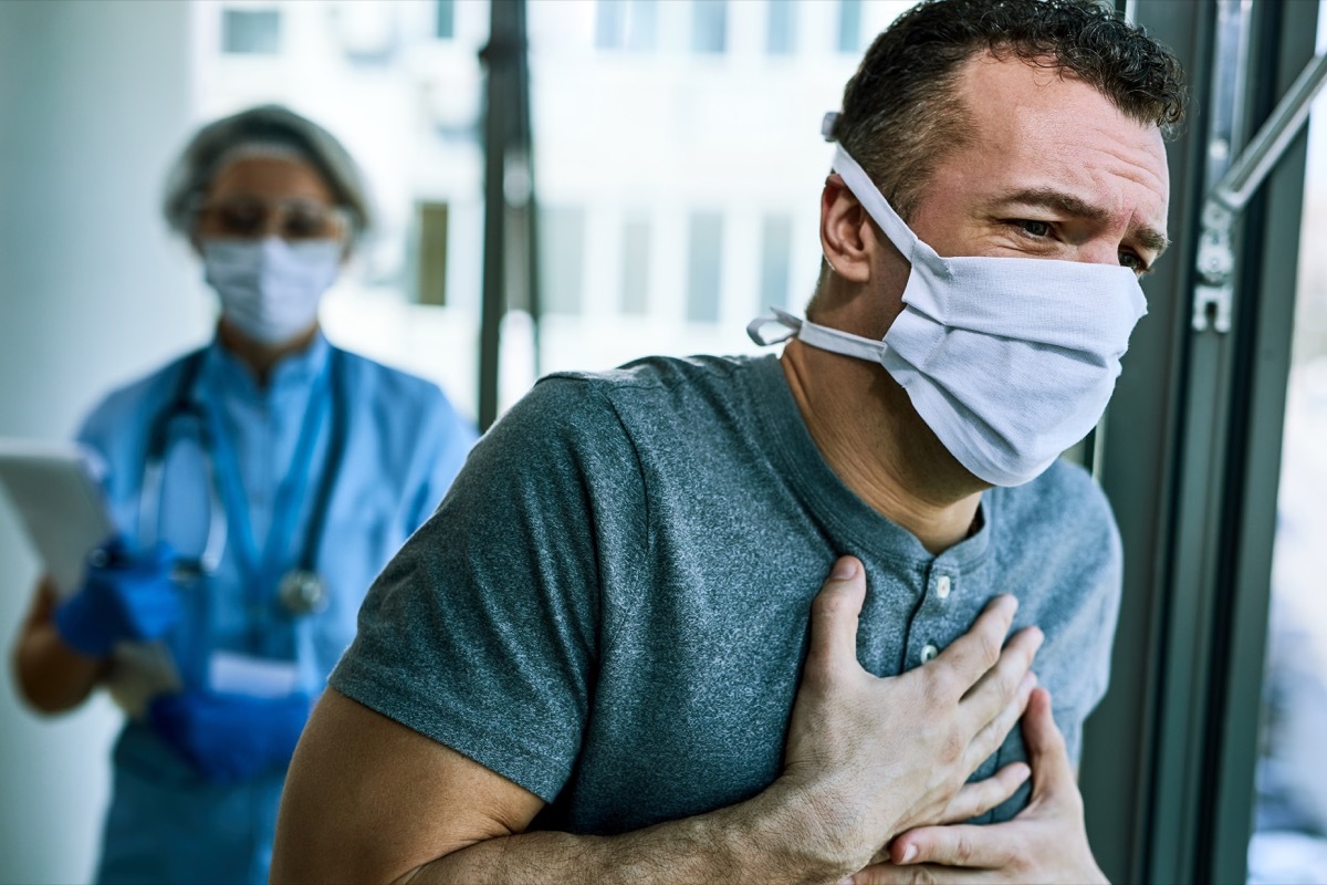 Male patient wearing face mask and feeling chest pain while being at the hospital during coronavirus epidemic. Healthcare worker is in the background.