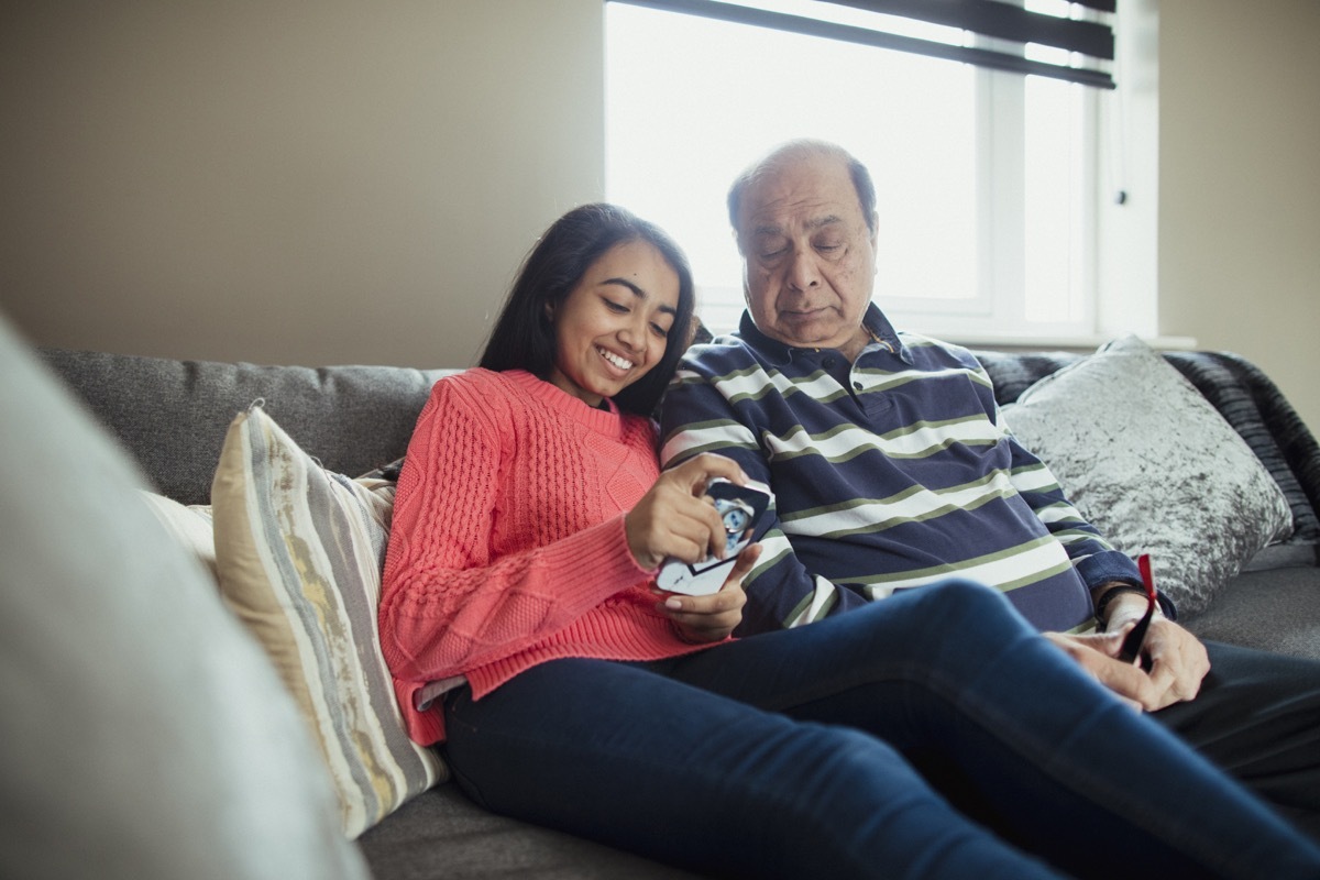 A front-view shot of a grandfather sitting down on a sofa with his granddaughter, the young granddaughter is using her phone to play an online game.