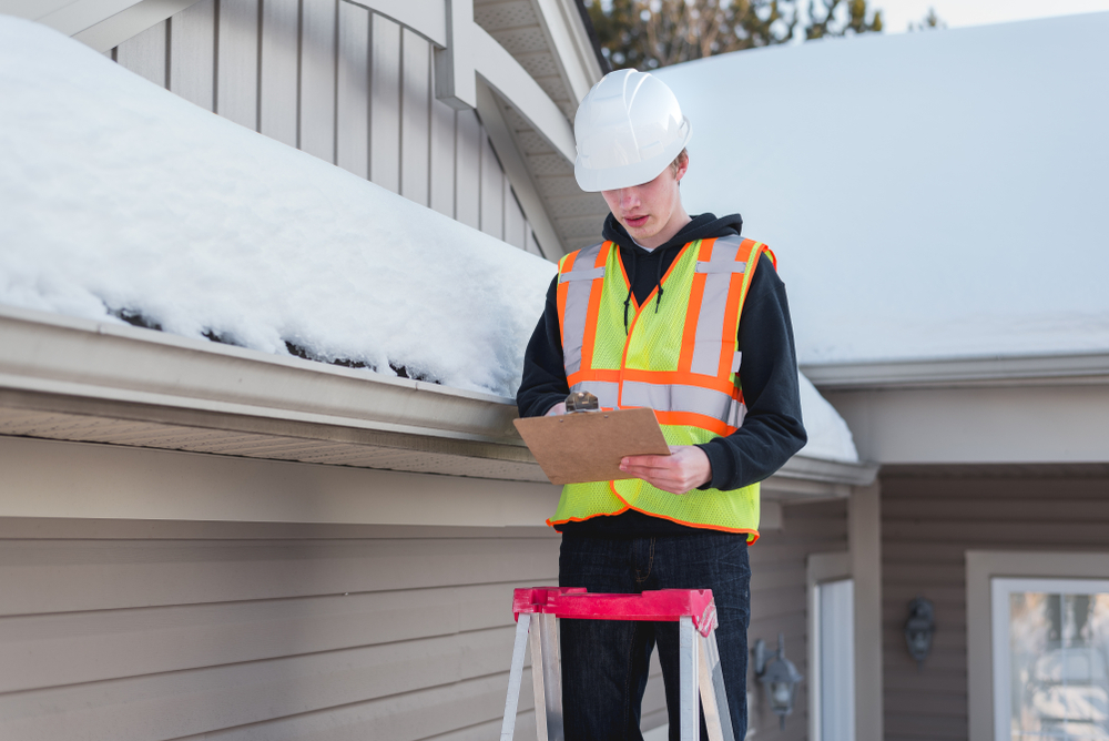 A home inspector on a ladder checking a roof covered in snow