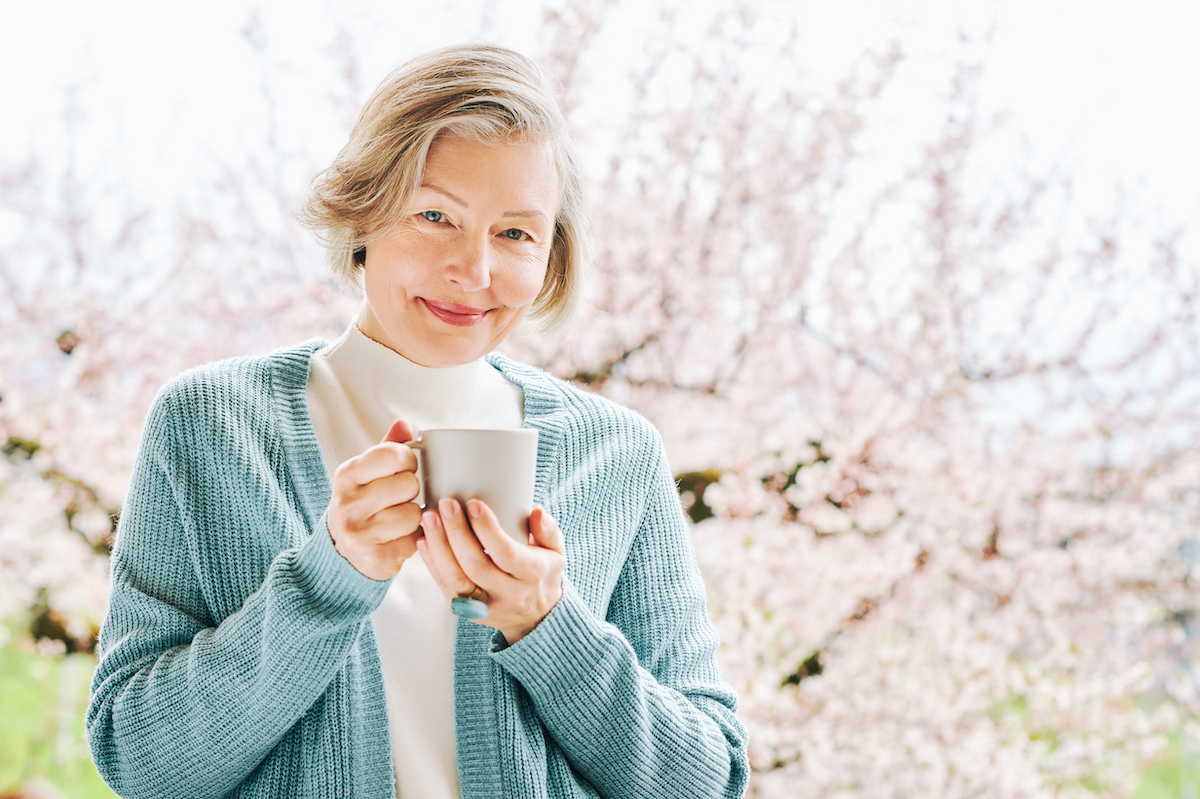 An elegant older woman holding a mug and standing against a background of cherry blossom trees.