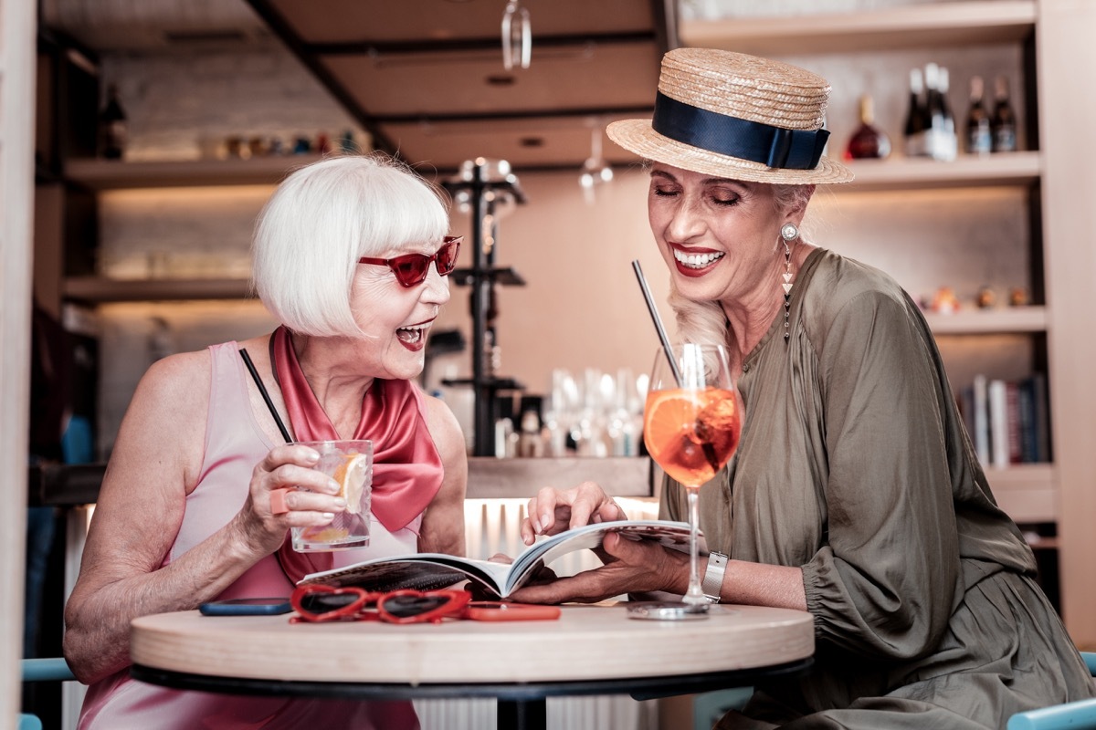 Two older women have drinks and read book together, one wears a hat indoors, long marriage tips