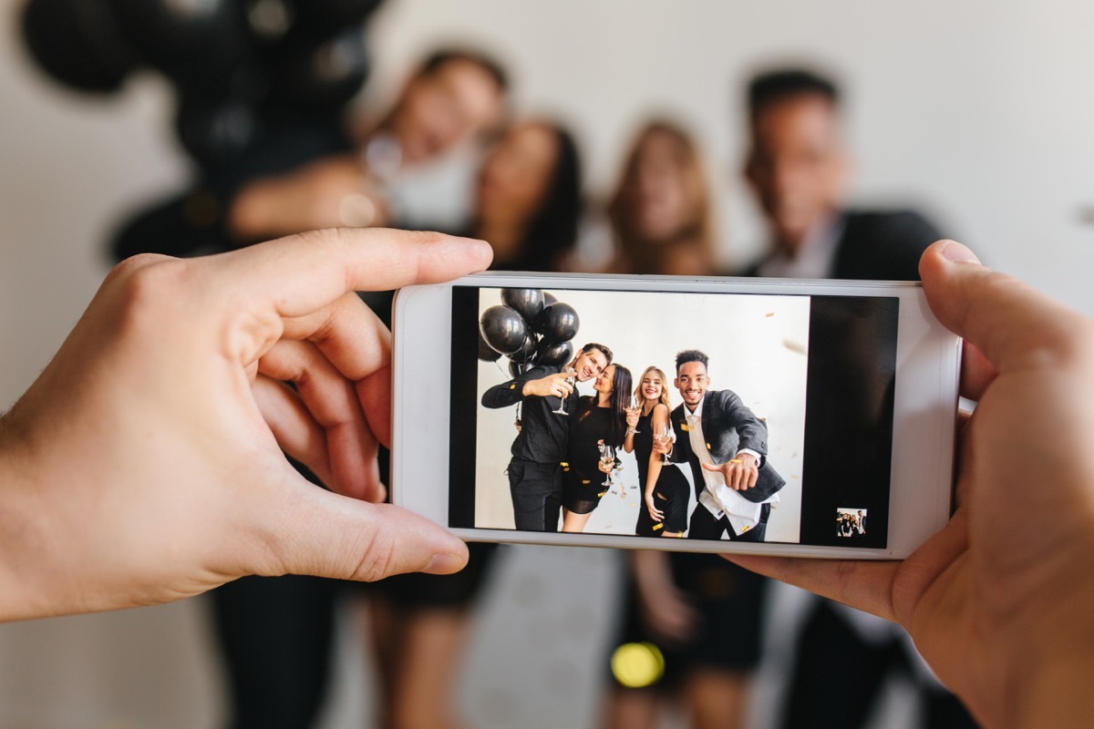 Blur portrait of blissful young people posing with balloons at birthday party with hands holding phone on foreground. Man taking photo of friends celebrating something and using smartpone. - Image