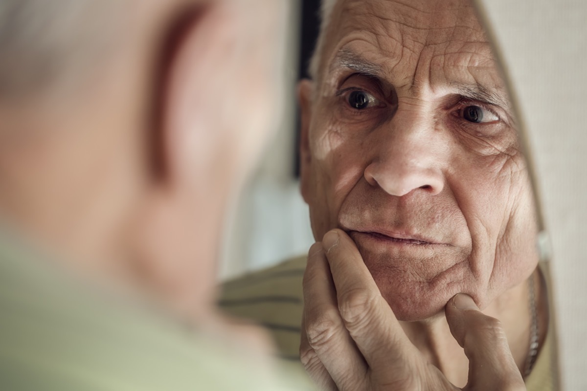 Sad senior man looking in bathroom mirror and touch his face skin with fingers. Concept grow old.