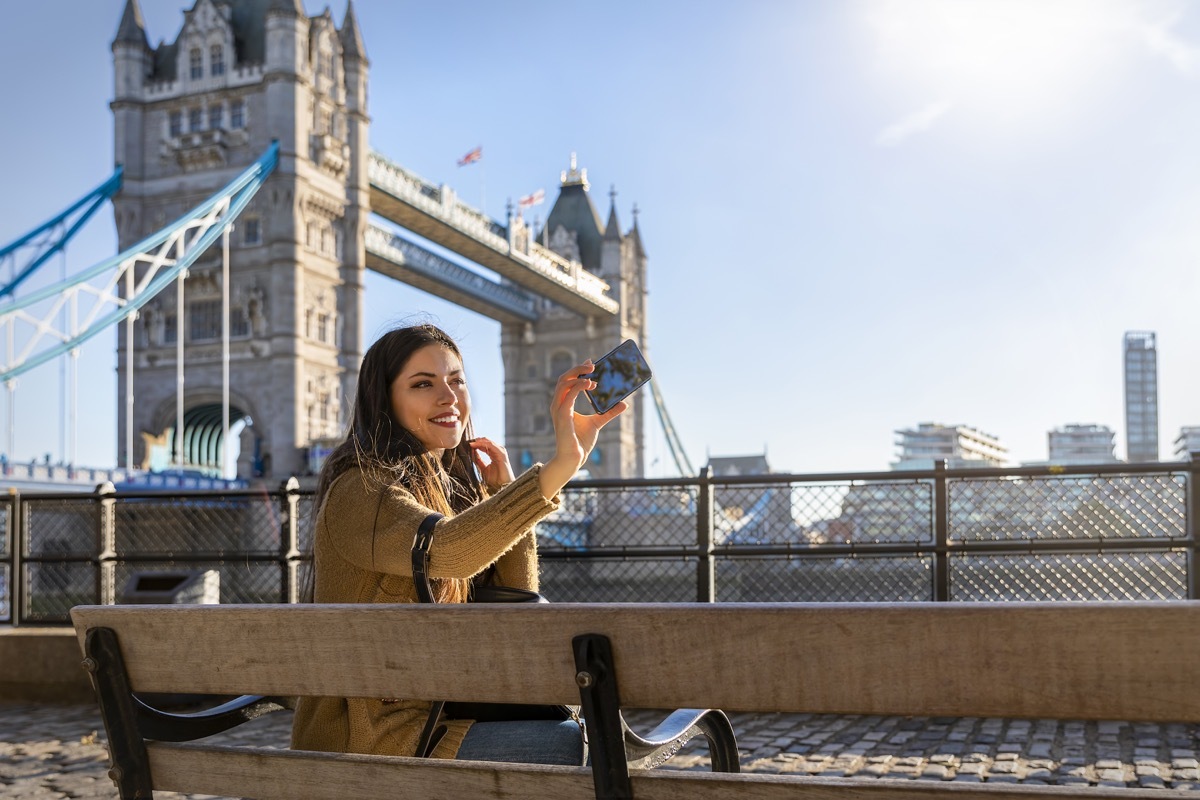 young woman sitting on bench and taking a selfie with Tower Bridge in the background in London on a sunny day