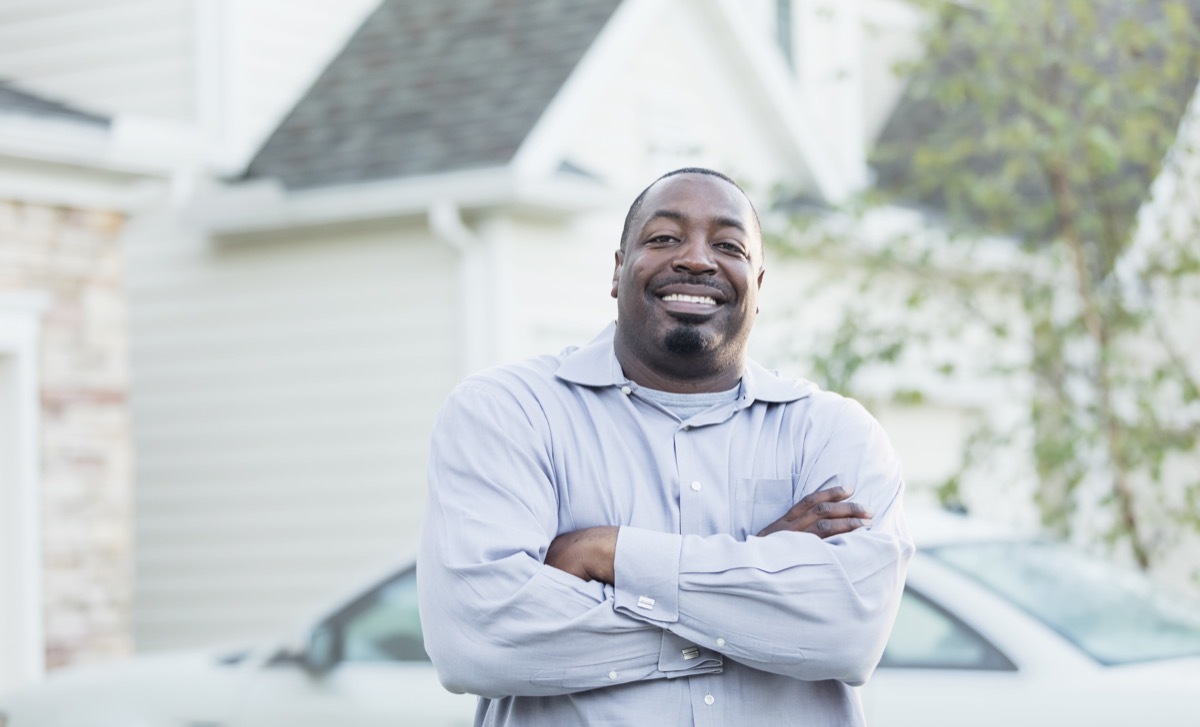 A mature African-American man in his 40s standing outdoors by the driveway of his home, smiling at the camera with his arms crossed.