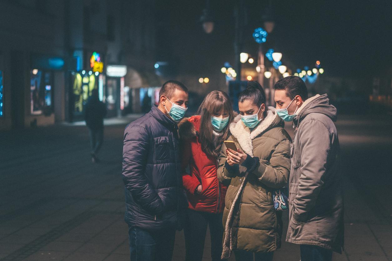 A group of young friends wearing face masks look at a smartphone in one of their hands.