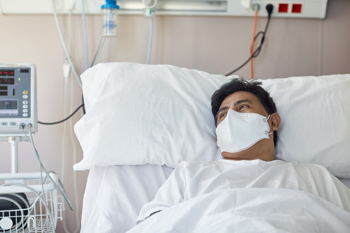 Portrait of male patient in early 40s looking away from camera while lying in hospital bed wearing protective face mask and recovering from coronavirus.