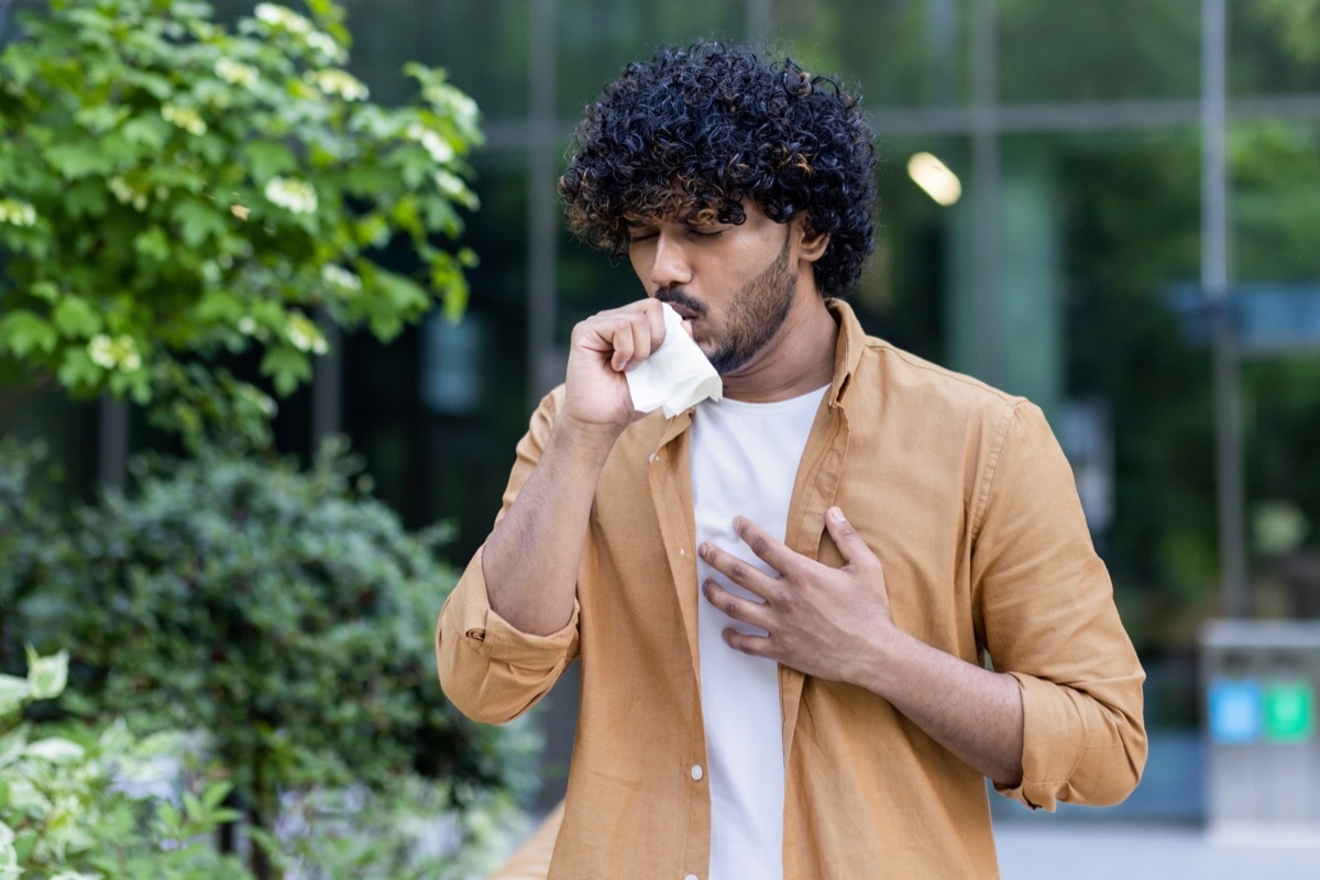 A young man is standing outside on the street and coughing.