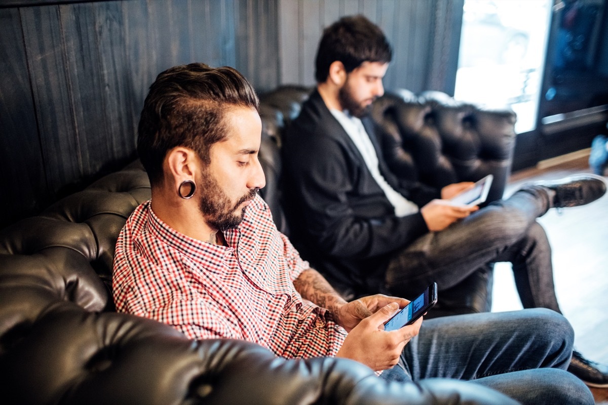 Mid adult man using mobile phone on sofa in hair salon. Male clients are waiting at barber shop. They are wearing casuals.