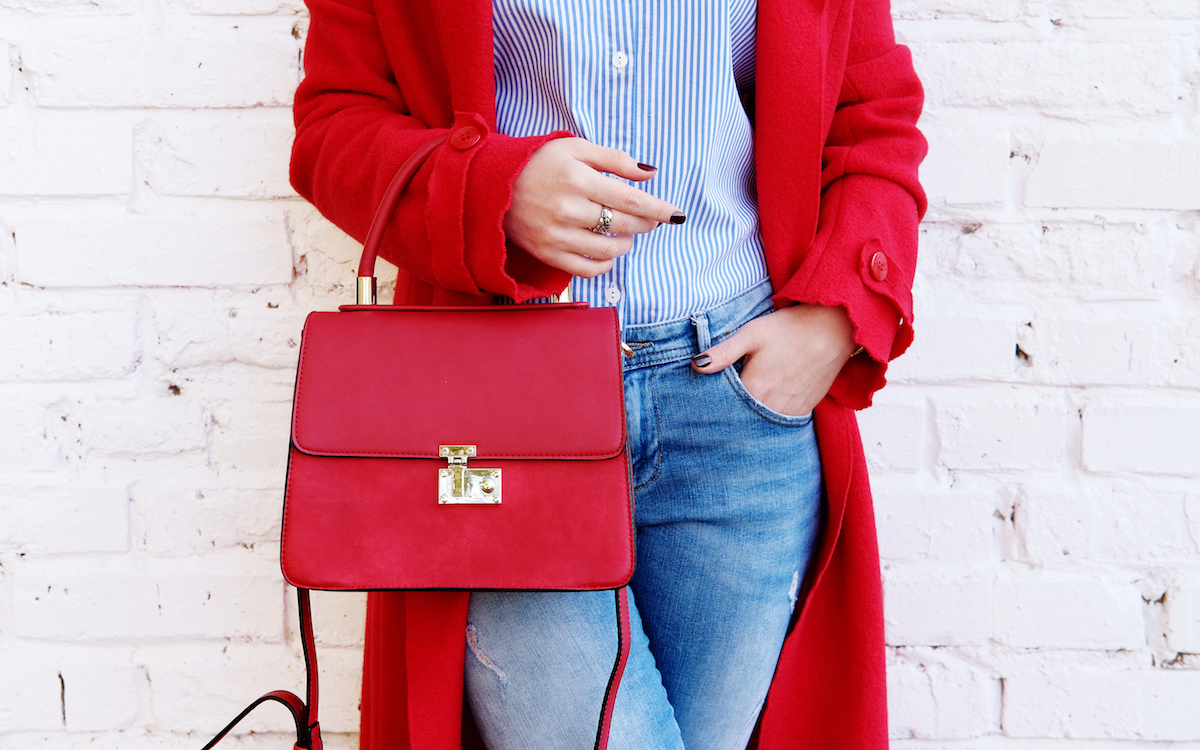 Closeup of red small bag in hand of woman. Fall spring fashion outfit red coat and trendy blue jeans