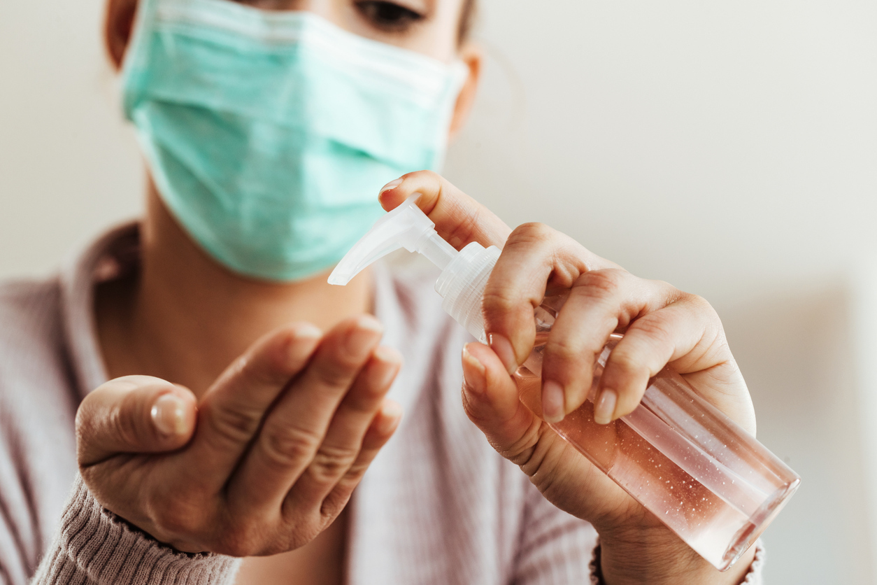 A woman wearing a sweater and a face mask uses a pump bottle with hand sanitizer.