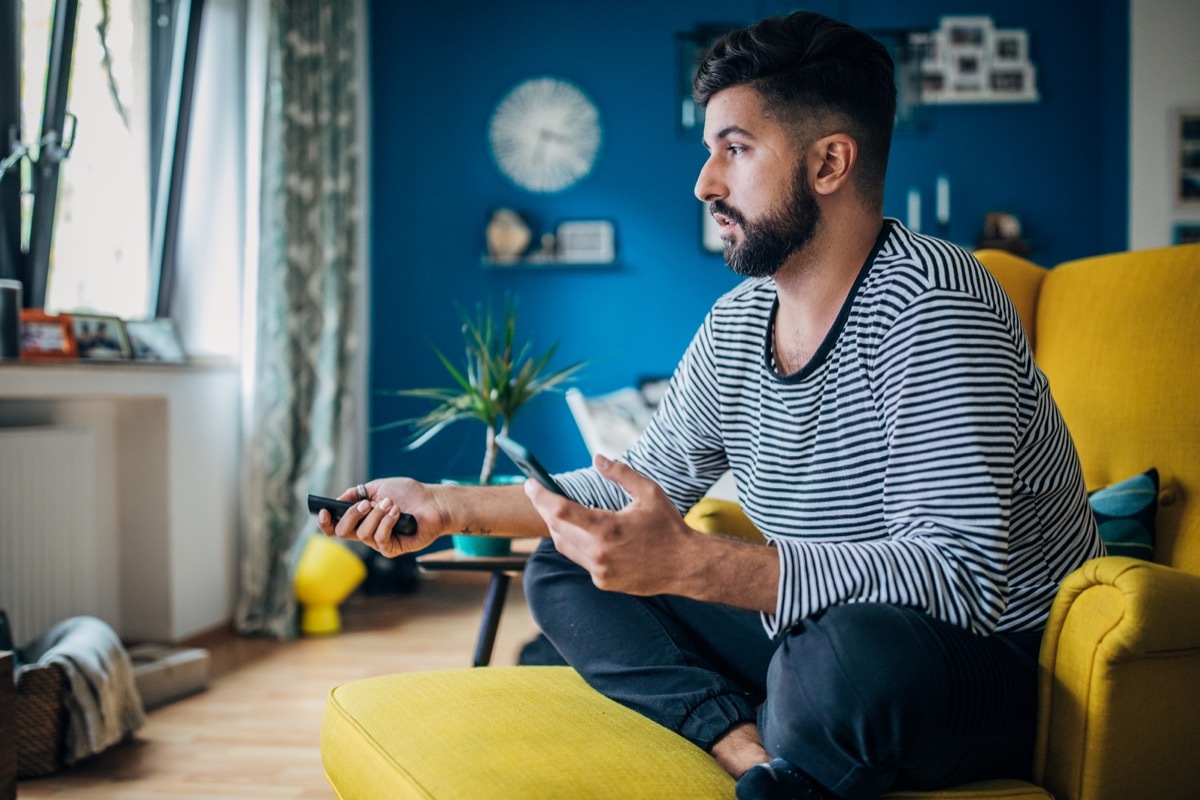 Man sitting home in his armchair, using phone and changing channels