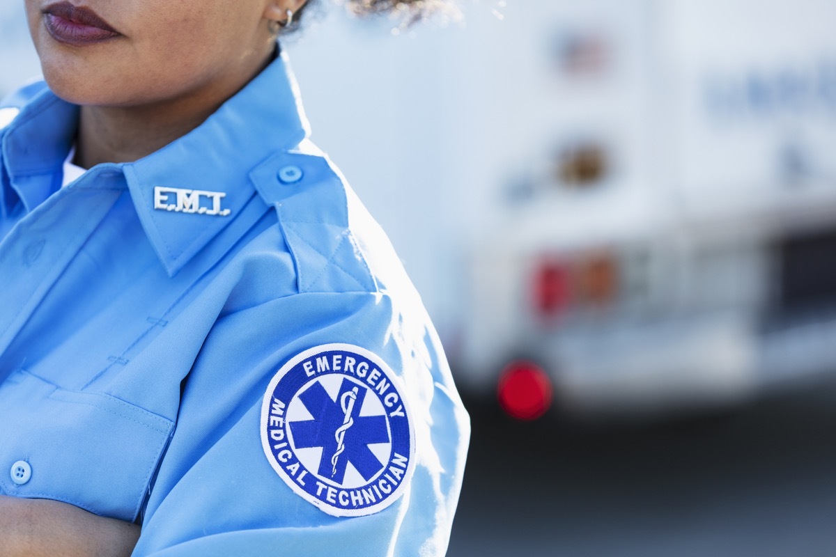 Cropped view of a female paramedic standing with arms crossed in front of an ambulance. She is a mid adult woman in her 30s, mixed race Hispanic and Pacific Islander. The focus is on the generic EMT patch on her sleeve at the shoulder.